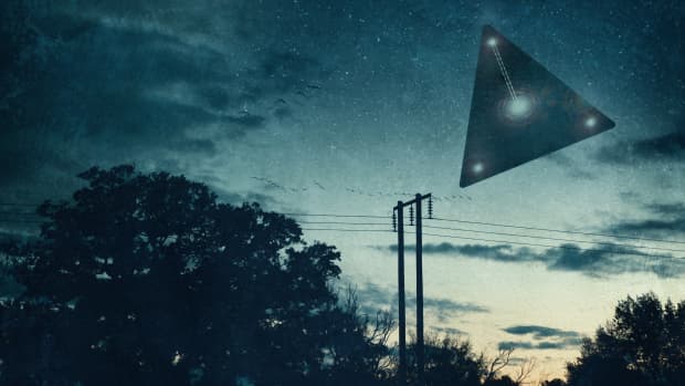 Black, triangle shaped UFO with a light in each corner and one in the center flying above telephone lines at dusk.