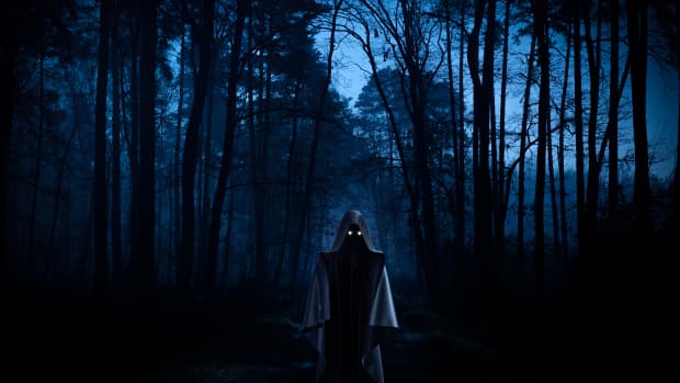 a hooded creepy figure stands in the darkened woods