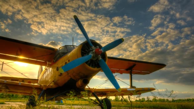 An old yellow bi-plane with a bright blue propeller sits in a field in front of a glorious sunset