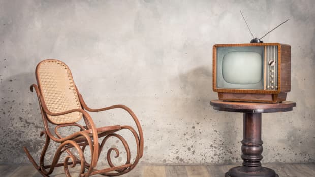 Retro old outdated TV receiver from circa 1950s on wooden table and aged rocking chair in front of textured concrete wall background.