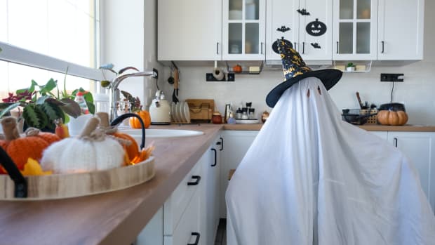 Child dressed as a ghost in a white sheet with two eye-holes and a pointed witch's hat standing in the kitchen.