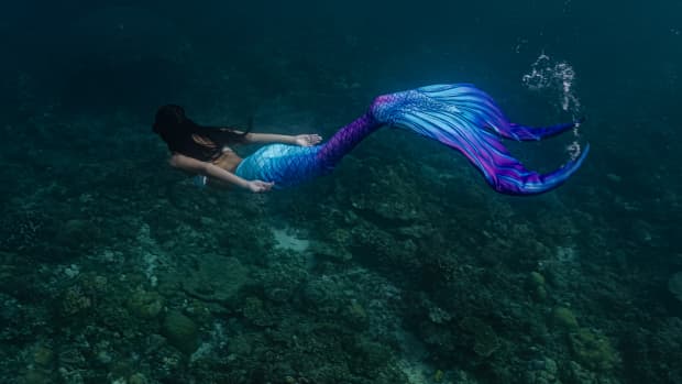 A woman in a realistic mermaid costume with a turquoise and purple tail swims though a grotto