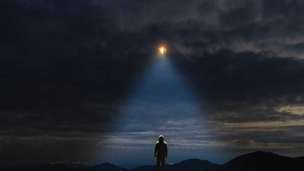UFO in a cloudy sky shines light down on a man standing on the ground