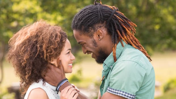 a Black couple with natural and dreadlocked hair faces each other, his hands on her face. both smiling broadly