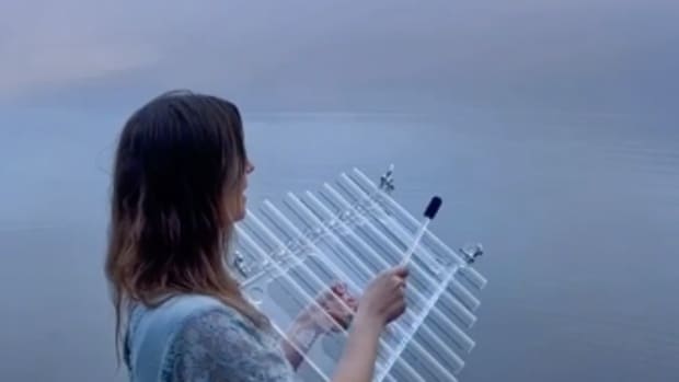 A white woman with brown hair wades in a body of water underneath a periwinkle and pink sky, singing as she plays and instrument that looks like a glass xylophone.