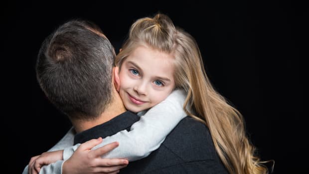 A blond girl child hugs an older man. She is facing the camera and he is facing away.