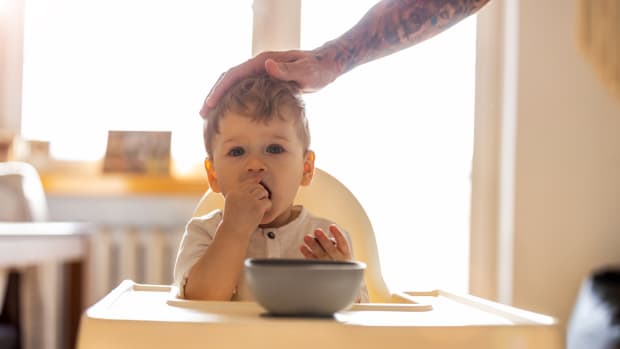 A tattooed man's hand caresses the head of an adorable white toddler boy sitting in a high chair.