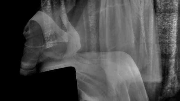 a ghostly figure in historical dress seems to stand up from her body. Picture is black and white.