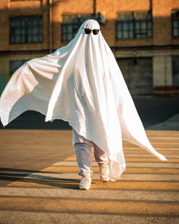 Person wearing a sheet like a ghost and sunglasses skips down a sunny street.