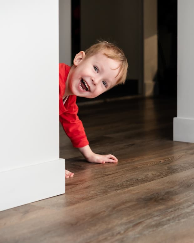 A white male toddler in a red shirt peeks out from behind a doorway, smiling.