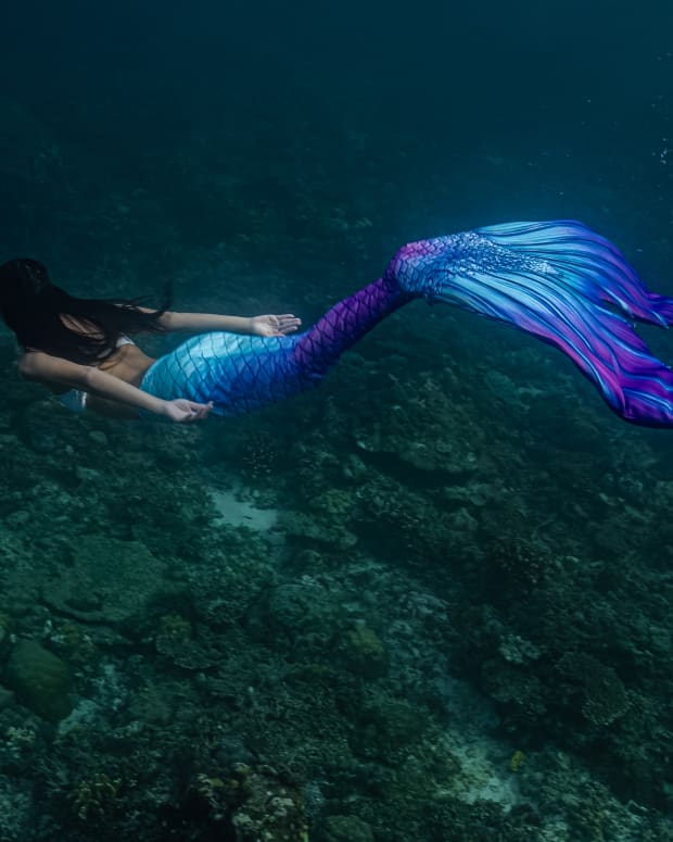 A woman in a realistic mermaid costume with a turquoise and purple tail swims though a grotto