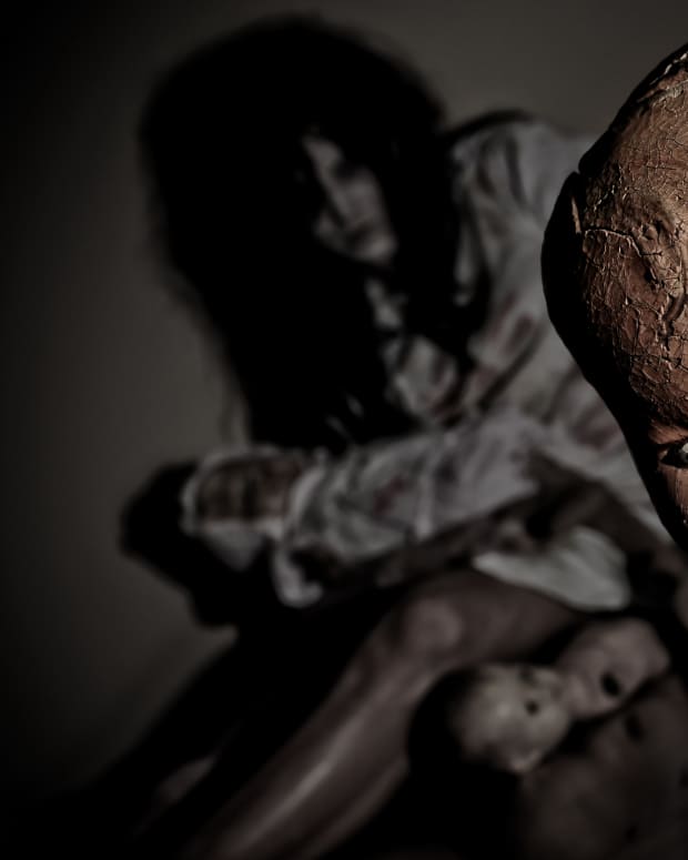 A cracked old doll looms in the foreground while a creepy female figure holding yet another scary doll lurks in behind it