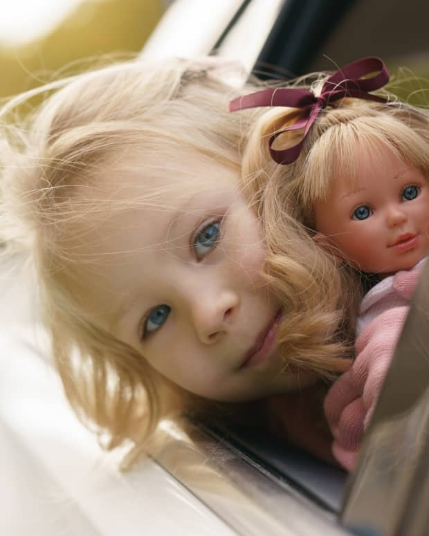 Little blonde girl leans out of car window holding a little blonde girl doll that looks just like her.