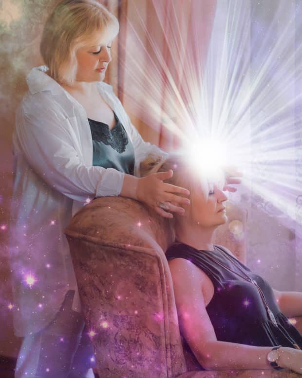 A woman stands behind another, seated woman, her hands on the seated woman's head. a bright, shimmering light appears to come from the seated woman's forehead.