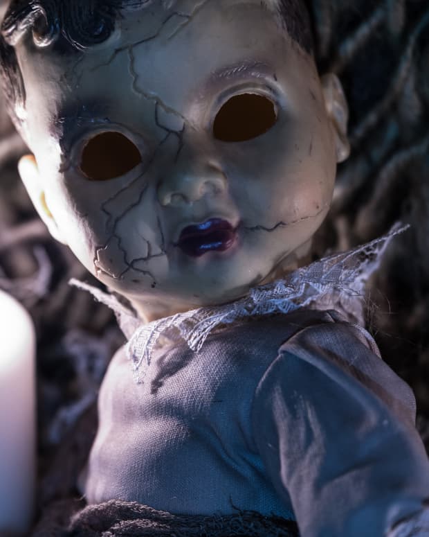 Creepy hollow-eyed doll looks at viewer while a candle burns a ghostly flame