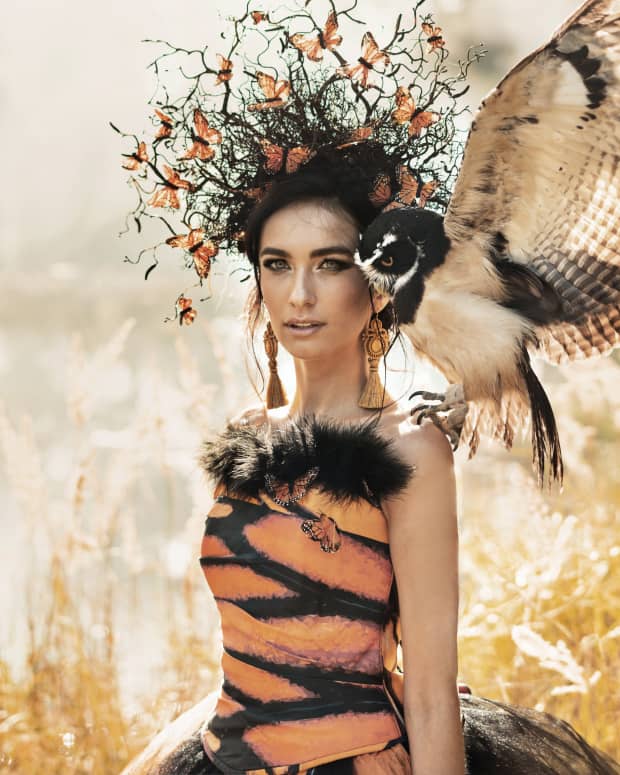 a white woman with dark hair in an ornate butterfly and leaves head dress looks at the camera. An owl sitting on her shoulder spreads its wings.