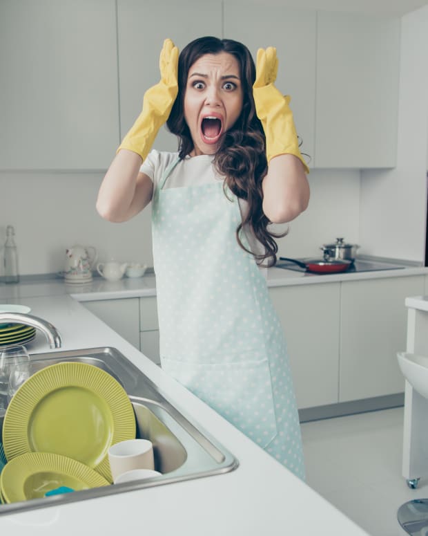 A white woman with dark hair stands in a pristine kitchen looking horrified, her dish-gloved hands raised to her head. A plate of clean dishes are in the sink in front of her.