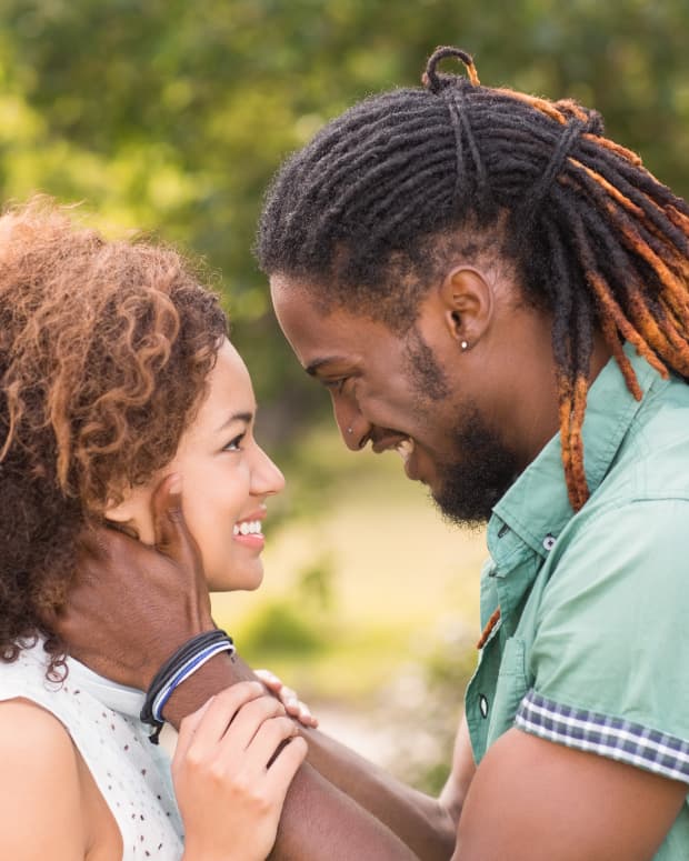a Black couple with natural and dreadlocked hair faces each other, his hands on her face. both smiling broadly