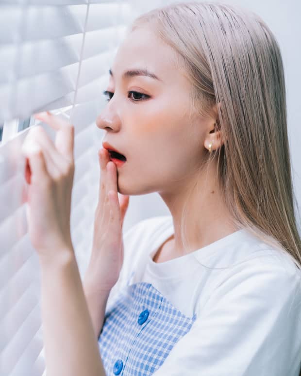 A young Asian woman with bleached hair peeks between blinds to look out of a window and brings her hand to her mouth in surprise