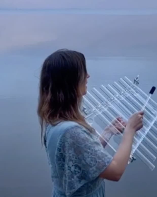 A white woman with brown hair wades in a body of water underneath a periwinkle and pink sky, singing as she plays and instrument that looks like a glass xylophone.