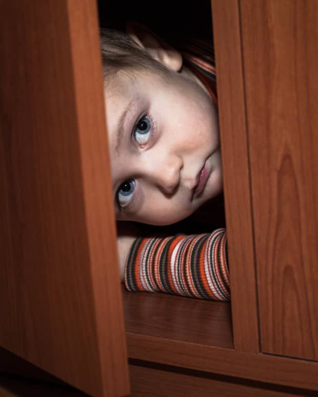 a white toddler boy peeks fearfully out of a partly open cabinet.