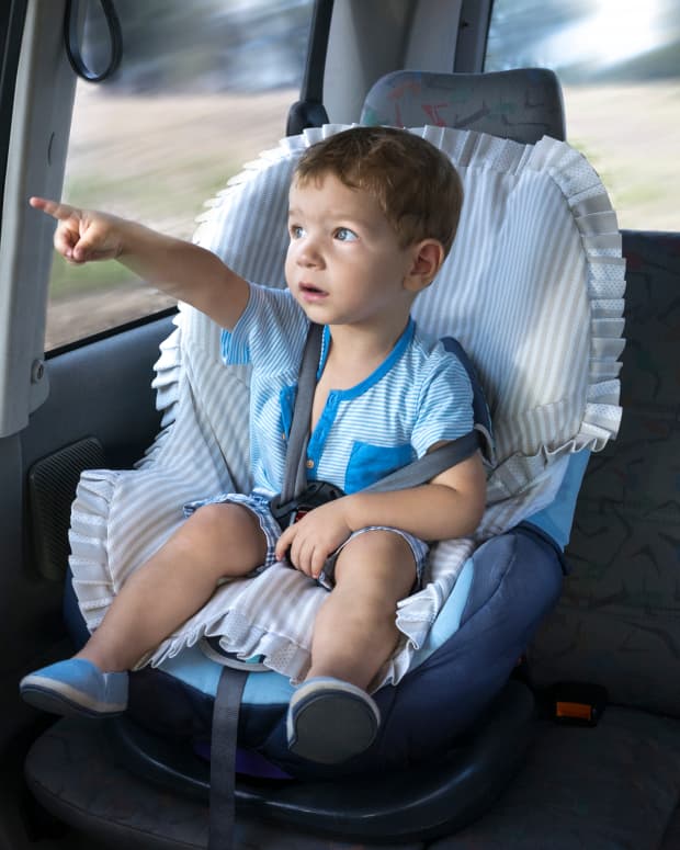 A toddler child in a car seat points out the window with a worried look on his face.