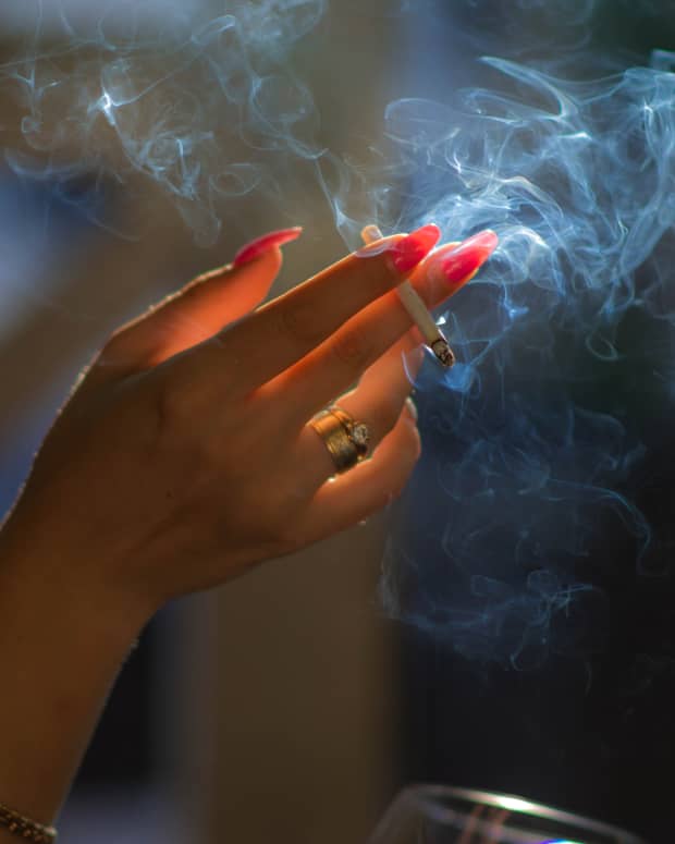A white woman's hand with long red fingernails waves in the air, holding a cigarette in her fingers, while smoke is eerily backlit.