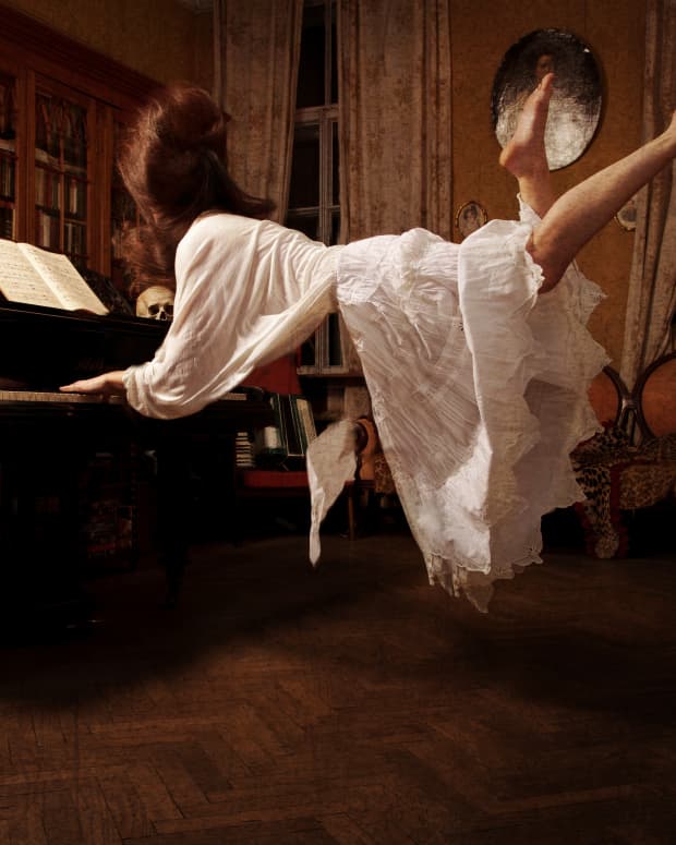 A white woman with a bouffant red hairdo and an ethereal white dress seems to float on air as she plays piano in an ornate and goth room.
