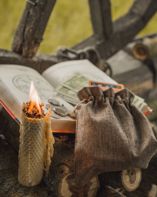a candle and a spell book and a bag of tarot cards crossed with money and coins.