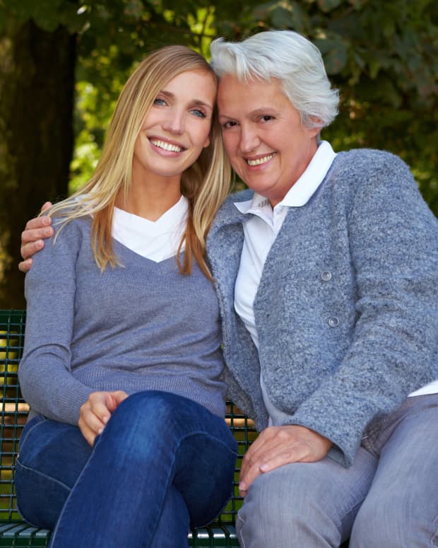 An old woman and young woman with their arms around each other smile at the camera from a bench