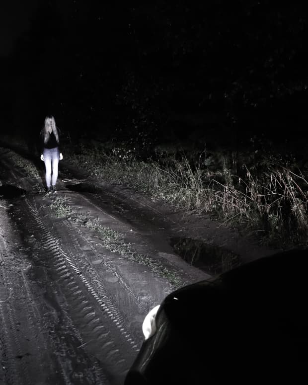a girl stands alone in a dark wooded road under the glare of headlights