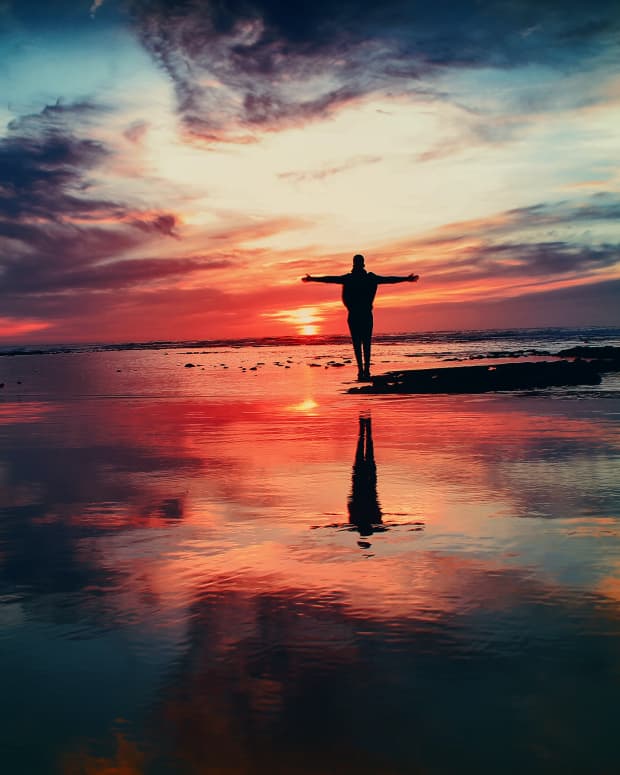 A silhouette of a man standing with his hands outstretched on a rock over water under a glorious sky