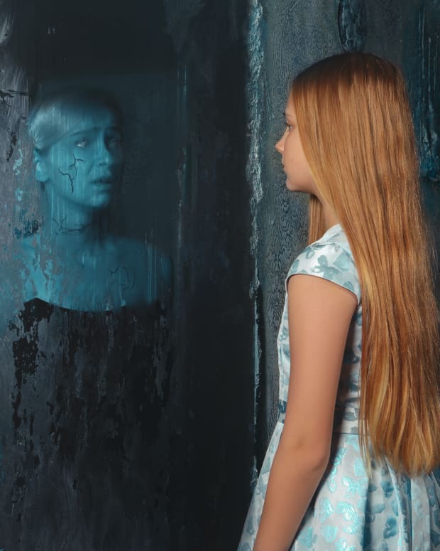 a white girl with long blonde hair looks into a mirror in the ruins of a house. a ghostly blue apparition looks fearfully back out.