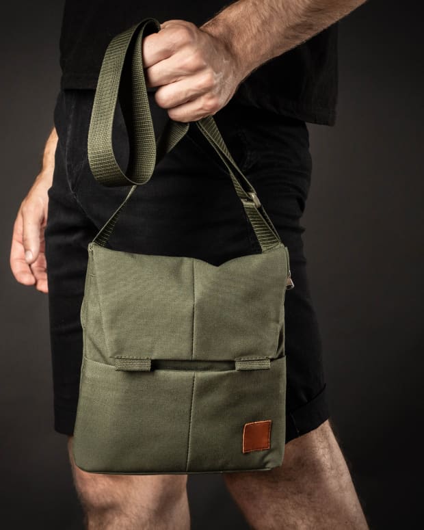 A white man wearing all black hold a khaki shoulder bag by the strap