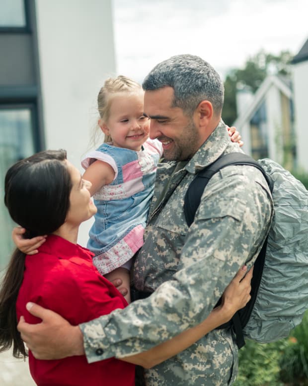 a man in camo fatigues holds a child and embraces his wife outside their home.