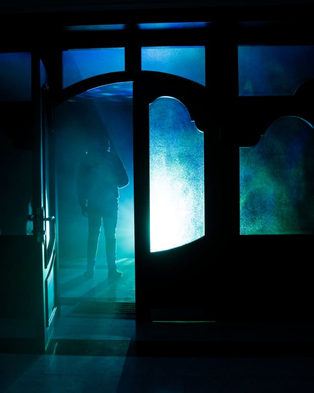 a shadowy silhouette stands in the threshold of a door, with a foggy blue-green light filling up the doorway and surrounding windows.