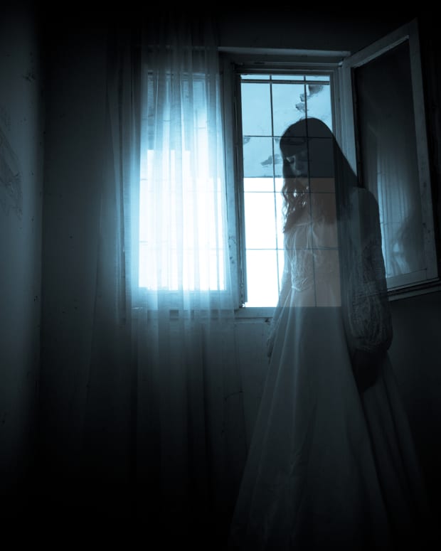 a semi-transparent woman in a long white gown stands silent and sad in front of a lit window in a dark room.