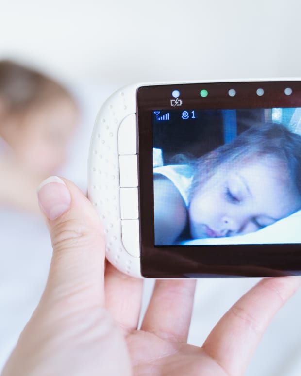 a close up on a hand holding a video baby monitor device showing a screen of a sleeping white toddler girl. In the background is the blurry image of presumably the same child, asleep in bed.