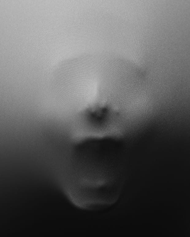 a screaming face presses through a piece of white or gray fabric
