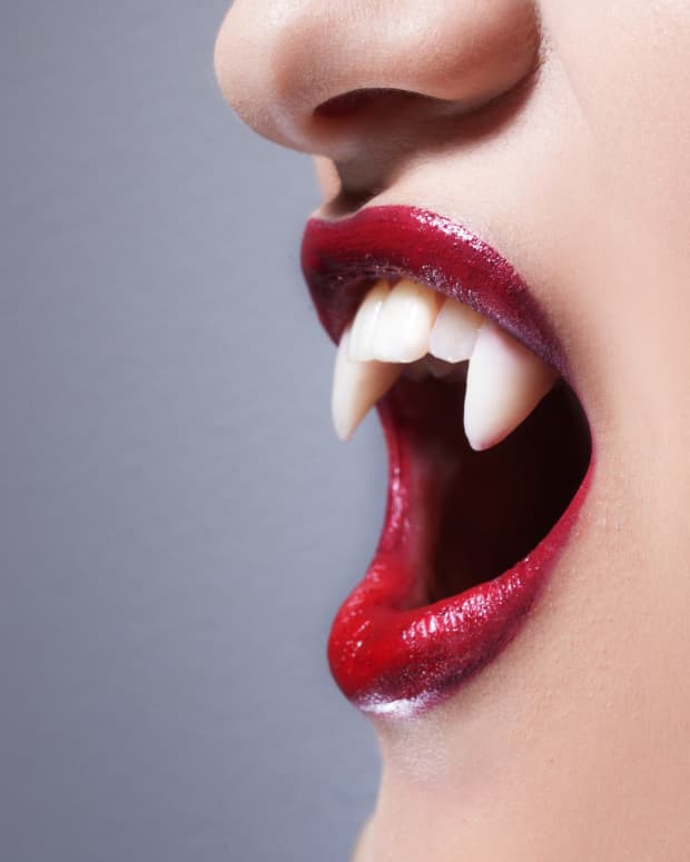 a close up on the lower half of a woman's face in profile. She is wearing dark red lipstick and has very realistic looking vampire fangs