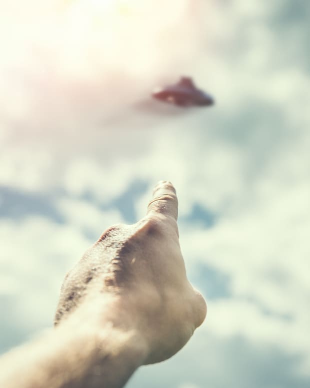 a man's hand points at an out of focus UFO in the sky.