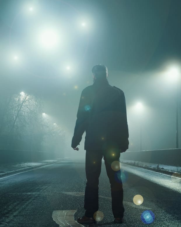 orbs shine in a dark sky with a man in the foreground wearing long pants and coat, looking up at them.