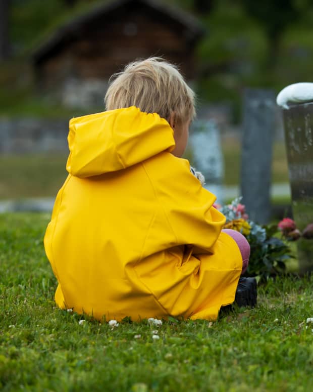 a little blonde white boy in a yellow raincoat sits in front of a gravestone with his back to the photographer