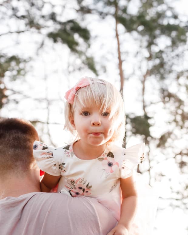 A man walks through the woods with a little girl picked up and looking at the camera over his shoulder