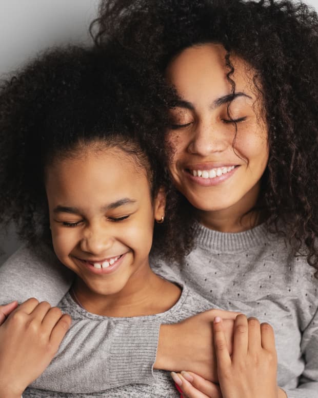 a light skinned black woman with natural hair hugs her daughter from behind. they are both smiling with their eyes closed.
