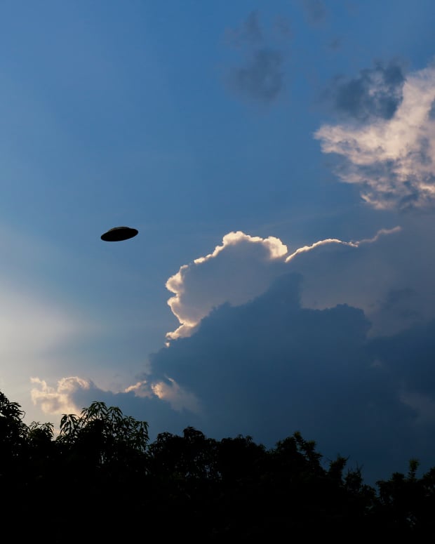 a strange flying saucer shaped object floats in a cloudy sky over some trees.