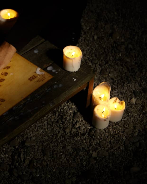 Three pairs of hands on a planchette our a Ouija board, with candles burning around it.