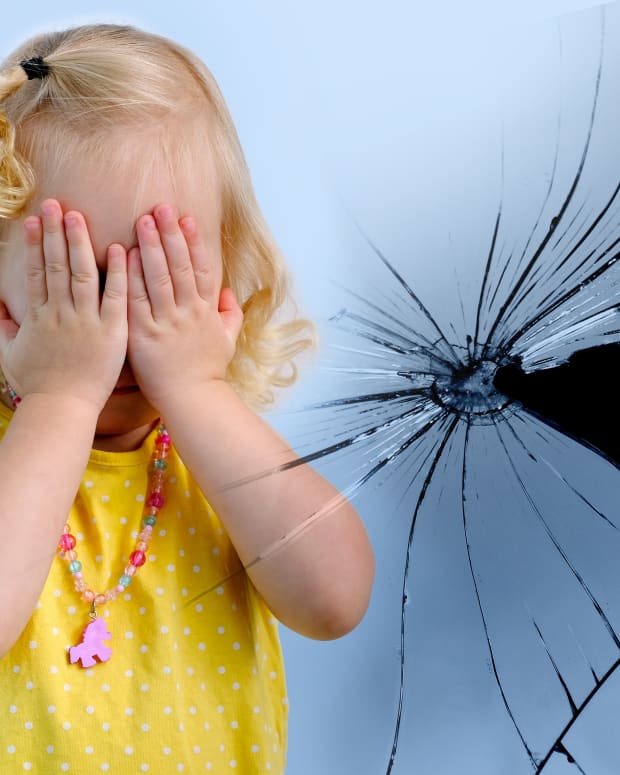 a blonde toddler holds her eyes over her hands in fright near a broken mirror
