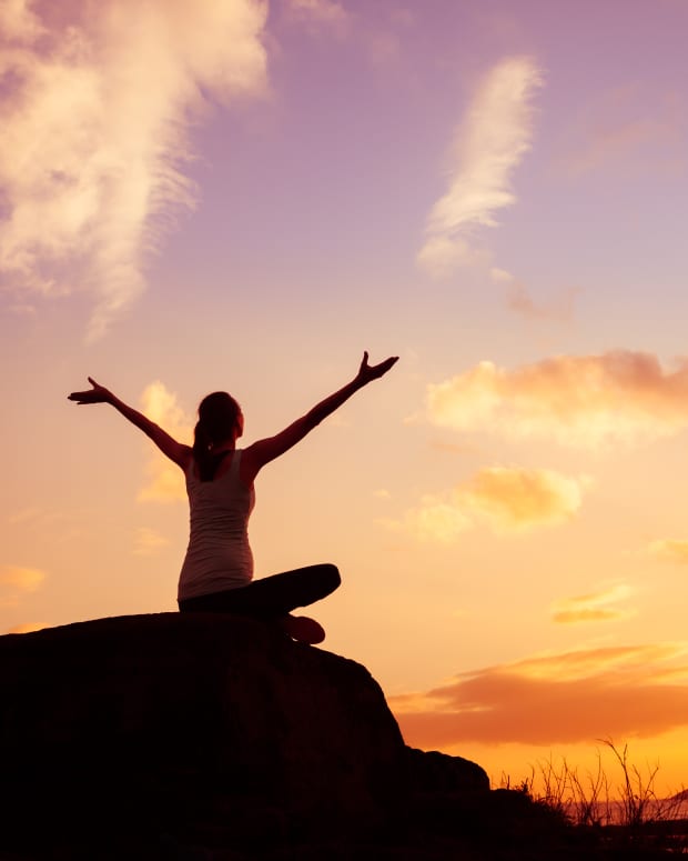a woman sits cross legged on a boulder with her back to the camera and welcomes a glorious sunrise with her arms held wide.