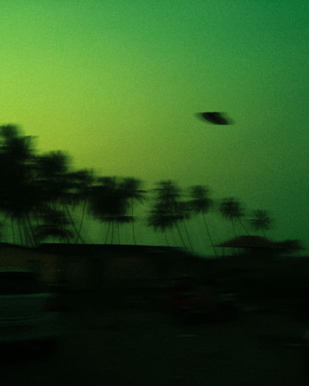 a dark UFO hovers in a blurry green sky over a parking lot lined with palm trees